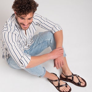 Pu&ograve; includere: Un uomo con una camicia a righe e jeans blu &egrave; seduto con le gambe incrociate, indossando sandali di cuoio marroni con pi&ugrave; cinturini.