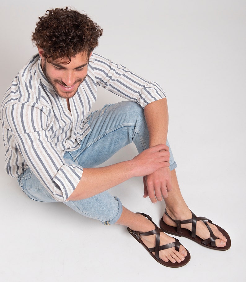 Pu&ograve; includere: Un uomo con una camicia a righe e jeans blu &egrave; seduto con le gambe incrociate, indossando sandali di cuoio marroni con pi&ugrave; cinturini.