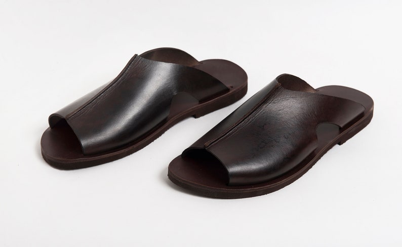 May include: A pair of dark brown leather sandals. The sandals have a simple design with a single strap across the top of the foot and a dark brown sole. The sandals are made of smooth leather and appear to be new.