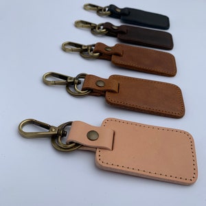 May include: A collection of five leather keychains in various shades of brown and black. Each keychain features a rectangular leather tag with stitched edges and a metal clasp. The keychains are arranged in a row, showcasing the different colors and textures.
