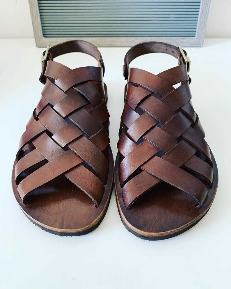 Puede incluir: Un par de sandalias de cuero marr&oacute;n con un dise&ntilde;o tejido. Las sandalias tienen un cierre de hebilla y un dedo del pie abierto.