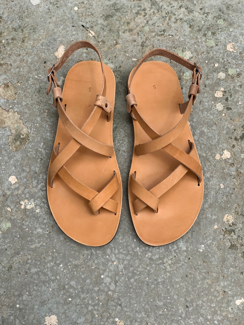 Pu&ograve; includere: Un paio di sandali in pelle color cuoio con cinturini incrociati e chiusura a fibbia.