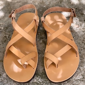 Pu&ograve; includere: Un paio di sandali in pelle color cuoio con cinturini incrociati e chiusura a fibbia.