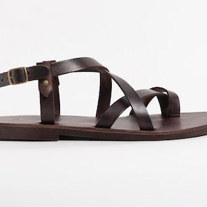 Pu&ograve; includere: Sandali in pelle marrone con un design a cinturino incrociato e una fibbia regolabile alla caviglia.