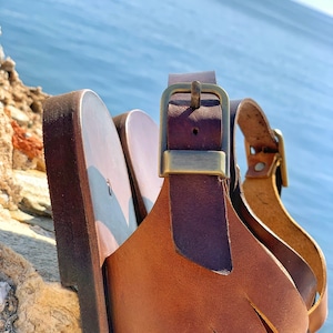 Puede incluir: Primer plano de una sandalia de cuero marr&oacute;n con una hebilla dorada. La sandalia tiene un dise&ntilde;o de correa tejida y est&aacute; sentada sobre una superficie rocosa.