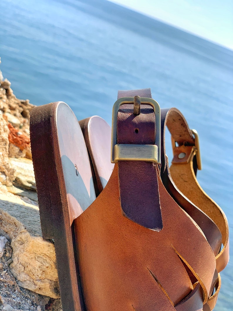 Puede incluir: Primer plano de una sandalia de cuero marr&oacute;n con una hebilla dorada. La sandalia tiene un dise&ntilde;o de correa tejida y est&aacute; sentada sobre una superficie rocosa.