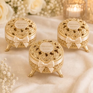 May include: Three round gold-toned metal trinket boxes with intricate filigree patterns, each featuring a cream-colored satin ribbon bow and a circular plaque engraved with the text EMMA & JAMES 06.20.2026, supported by three small decorative legs.