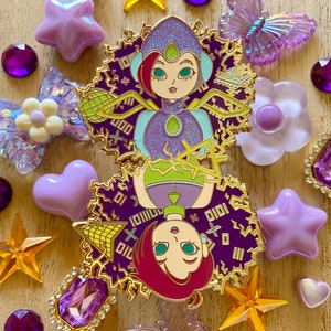 Pode incluir: Pin de esmalte dourado com duas mulheres de desenho animado com roupas roxas e verdes, rodeado por um design roxo e dourado. O pin tem um acabamento brilhante.