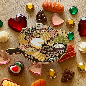 Puede incluir: Un pin de esmalte dorado que representa una tabla de charcutería con varios alimentos, entre ellos queso, fruta, galletas y carne. El pin está rodeado de pequeños corazones decorativos, gemas y otros pines con temática de comida.