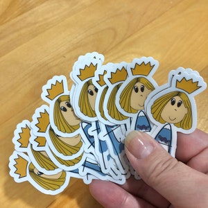 May include: A stack of stickers featuring a cartoon character with blonde hair, a crown, and a blue dress. The character is holding a surfboard with a mountain design.
