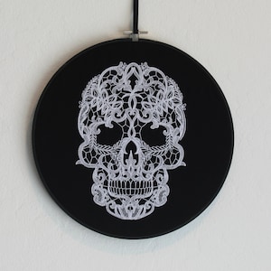 May include: A black embroidery hoop with a white lace skull design. The skull is detailed with intricate floral patterns.