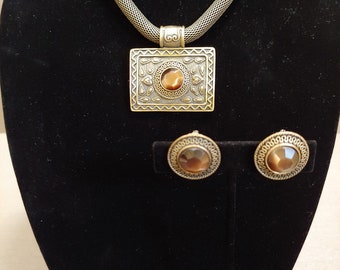 May include: A vintage-style jewelry set featuring a necklace and matching earrings. The necklace has a mesh chain and a rectangular pendant with an oval, brown stone. The earrings are round with matching stones. The metal has a bronze finish.