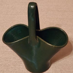 May include: A dark green ceramic basket with a curved handle. The basket has a flared, open design with a smooth, glossy finish. The interior of the basket is a darker shade of green.