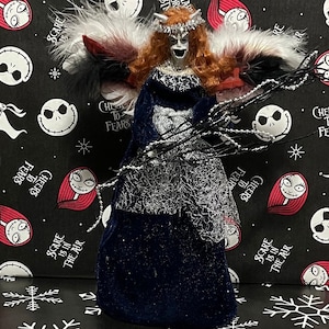 Op de afbeelding: Een blauwe en zilveren kerstboomtopper met een vrouwelijke figuur met rood haar, witte vleugels en een zwarte takvormige scepter. De figuur draagt een blauwe jurk met zilveren glitter en een zilveren kroon. De achtergrond is een zwarte stof met een herhalend patroon van Jack Skellington's gezicht en de tekst "Cheers to Fears" en "Scare is in the Air".