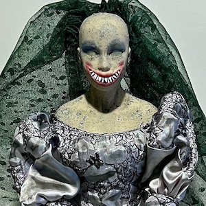May include: A porcelain doll with a green veil and a white lace dress. The doll has a painted face with a wide, toothy smile.