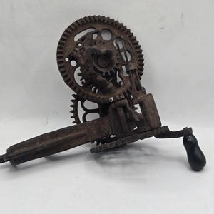 May include: A vintage hand-cranked drill with a heavy, rusted iron body and a large circular gear mechanism. The tool features a long, straight handle and a small, smooth black knob attached to the crank arm for manual operation and rotation.
