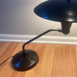 May include: A black desk lamp with a round, flat shade and a long, curved arm. The lamp is on a wooden surface.