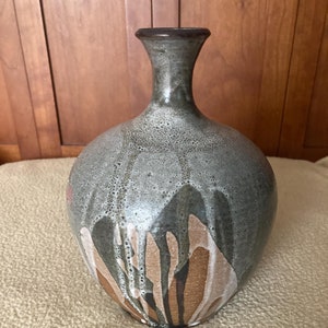 Puede incluir: Un jarrón de cerámica con un esmalte marrón, gris y blanco. El jarrón tiene un cuerpo redondo y un cuello estrecho.