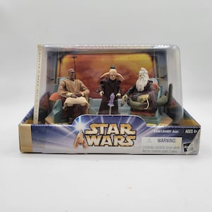 Peut inclure: Ensemble de figurines Star Wars, présenté dans un étui en plastique transparent. L'emballage affiche le logo Star Wars et le texte "Attack of the Clones". Les figurines représentent trois personnages dans une scène avec des chaises.