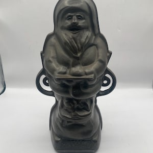 May include: A dark gray cast iron Santa Claus figure. The figure has a hooded cloak and holds a tray. The base of the figure has the text "HELLO DOLLIES". The figure has decorative scrollwork on the sides.