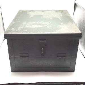 May include: A dark gray metal box with a rectangular shape and a hinged lid. The box has a lock and two latches on the front. The top of the box has a slightly weathered appearance.
