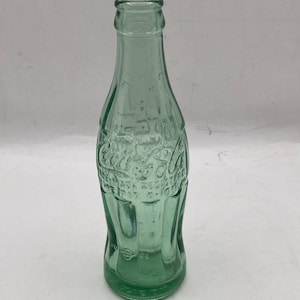 May include: A vintage light green glass Coca-Cola bottle with a contoured shape. The glass features embossed lettering that reads Coca-Cola and includes the text Marca Registrada. The bottle has a narrow neck and a wider base with vertical ridges.