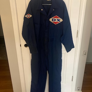 May include: Navy blue work jumpsuit with long sleeves and a collared neckline. The jumpsuit features two diamond-shaped patches with "DX" in white lettering and red and blue accents. The jumpsuit is hanging on a hanger.