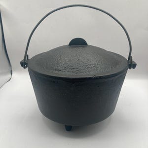 May include: A black cast iron Dutch oven with a lid and a handle. The pot has three small feet and a textured surface. The handle arches over the top of the pot.