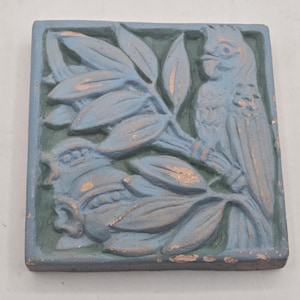 May include: A square ceramic tile featuring a relief design of a bird perched on a leafy branch. The tile is finished in a muted blue-gray glaze with dark green recessed areas and subtle copper-colored highlights on the raised surfaces.