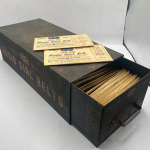 May include: A dark metal storage box labeled General Cement Radio Dial Belts with a pull-out drawer containing numerous tan paper envelopes. Two rectangular paper labels numbered 167 and 118 are placed on top of the box, each featuring the text Non-Slip Radio Dial Belt.