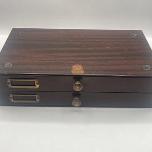 May include: A dark brown wooden box with two drawers. The drawers have brass-coloured handles and label holders. The top of the box is a darker shade of brown with a wood grain pattern. A small, round emblem is on the top.