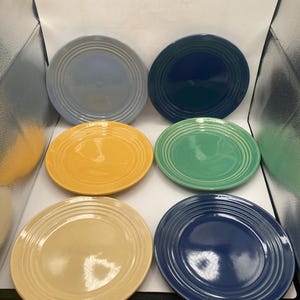 May include: Six ceramic plates in various colours: light blue, navy blue, yellow, green, and cream. Each plate has a circular design with concentric rings. The plates are arranged on a white surface.