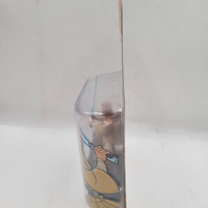 May include: A Star Wars action figure in a clear plastic package. The figure is of a Jedi with a blue lightsaber and tan robes. The packaging has a brown and orange color scheme.