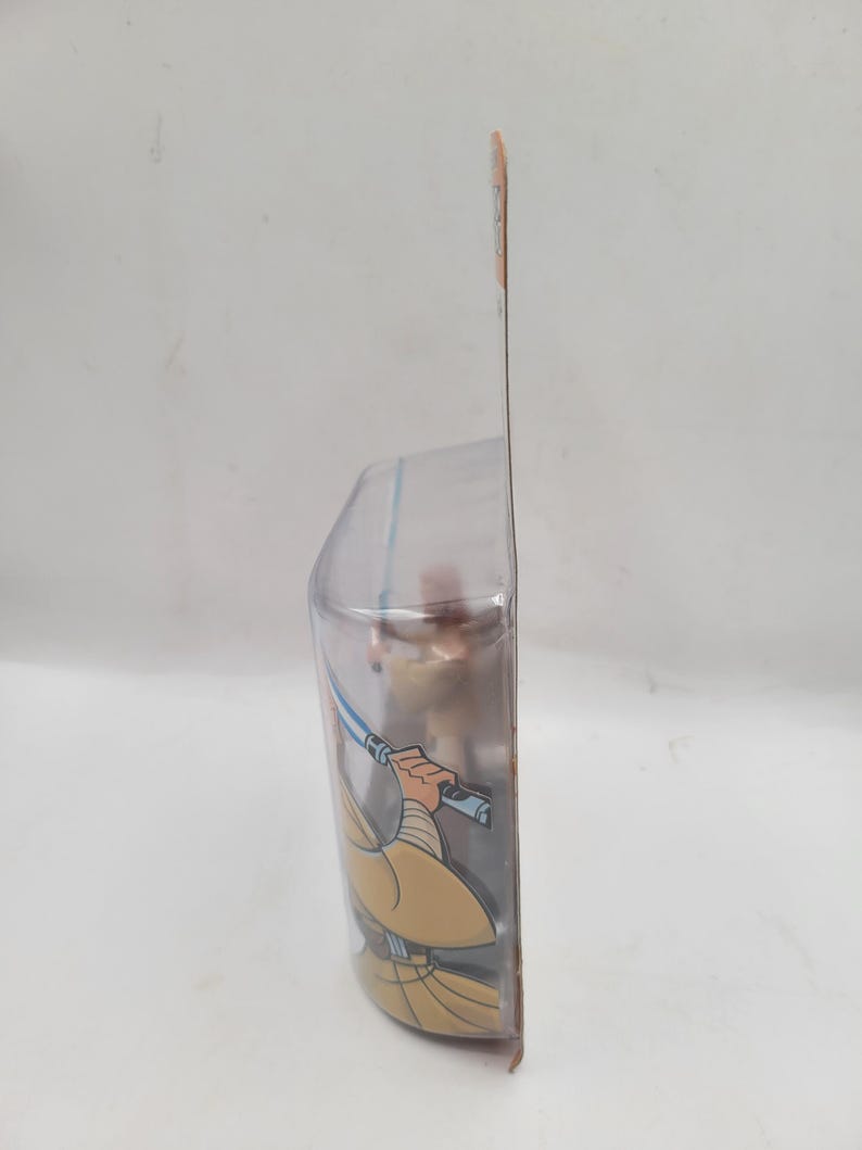 May include: A Star Wars action figure in a clear plastic package. The figure is of a Jedi with a blue lightsaber and tan robes. The packaging has a brown and orange color scheme.