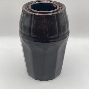May include: A dark brown, antique stoneware crock with a cylindrical shape and a slightly flared rim. The crock has vertical ribbing and a dark interior, suggesting it may have been used for storage. The surface has a glossy finish.