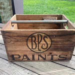 May include: A vintage wooden crate with the text "BPS PAINTS" in black lettering. The crate has a blue handle and a circular logo. The wood has a natural, aged appearance, suggesting it's an antique or a piece with a rustic aesthetic.