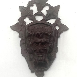 May include: A dark brown, ornate wall hanging featuring a sculpted face with a beard and leafy accents. The decorative piece has a detailed, antique appearance, with a symmetrical design and a textured surface. The overall shape is a stylized shield.