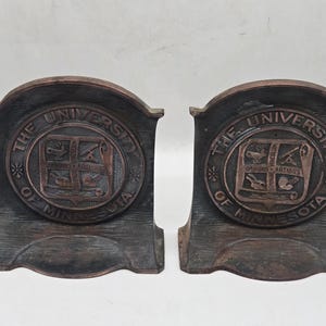 Könnte beinhalten: Ein Paar Buchstützen aus dunkel bronzefarbenem Metall mit einem kreisförmigen Emblem und der Aufschrift THE UNIVERSITY OF MINNESOTA. In der Mitte jedes Emblems befindet sich ein Wappen mit vier Quadranten, Symbolen und dem lateinischen Text OMNIBUS ARTIBUS.