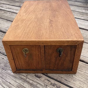 May include: A rectangular wooden box with two small drawers. The box is made of light brown wood with a visible wood grain. Each drawer has a small, ornate metal pull. The box is sitting on a wooden surface.
