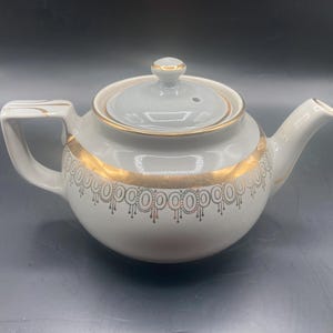 May include: White ceramic teapot with a light gray lid and gold trim. The teapot features a rounded body, a curved spout, and a handle. The gold trim includes a decorative pattern around the body. The lid has a small knob.