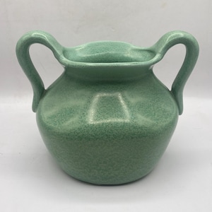 May include: A green ceramic vase with two curved handles. The vase has a speckled texture and a wide, open mouth. The handles curve upwards from the body of the vase. The overall shape is rounded, with a slightly flattened base. The vase is a muted green color.