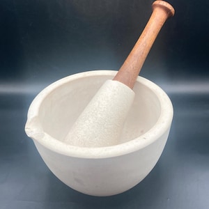 Puede incluir: Un juego de mortero y mano de mortero blancos. El mortero es un cuenco redondo con un pico, y la mano de mortero es de madera y piedra blanca. La mano de mortero está dentro del mortero.