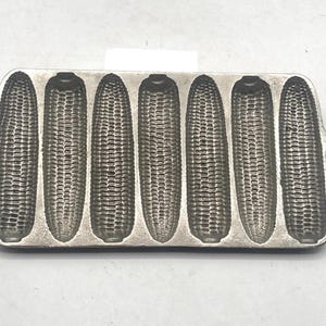 Op de afbeelding: Een gietijzeren maisbroodpan met zeven maïskolfvormen. De pan is rechthoekig met afgeronde randen en handvatten aan elk uiteinde. De vormen zijn gedetailleerd met de textuur van maïskorrels.