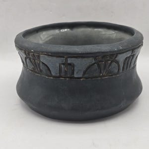 May include: A matte black ceramic bowl with a wide, rounded base and a slightly flared rim. The upper section features a decorative band with a repeating geometric pattern in a muted blue-grey glaze, while the interior has a smooth grey finish.