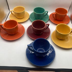 Pode incluir: Uma coleção de oito xícaras de chá e pires de cerâmica coloridos. As xícaras e os pires são em várias cores, incluindo amarelo, laranja, vermelho, verde-azulado, bordô e azul. Cada xícara tem uma alça e fica em um pires correspondente.
