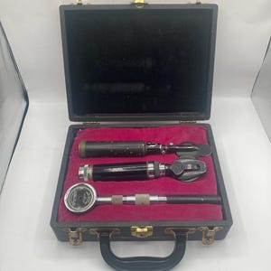 May include: A vintage medical instrument set in a black case with a handle. The case is open, revealing three instruments on a red velvet lining. The instruments include an ophthalmoscope and other tools.