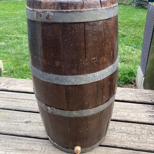 Könnte beinhalten: Ein verwittertes, dunkelbraunes Holzfass mit drei Metallbändern. Das Fass hat einen Zapfhahn in Bodennähe und steht auf einer Holzterrasse. Die Höhe des Fasses beträgt etwa 76 cm.