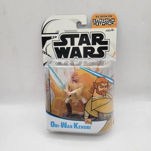 May include: Obi-Wan Kenobi action figure from Star Wars, in a Cartoon Network package. The figure is in a dynamic pose with a lightsaber, set against a fiery background. The packaging features the Star Wars logo and the text "As Seen On Cartoon Network."
