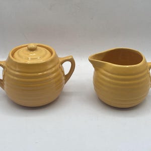 May include: A yellow ceramic sugar bowl with a lid and a matching creamer. Both pieces have a ribbed design and handles. The sugar bowl has a round shape, while the creamer has a spout for pouring.