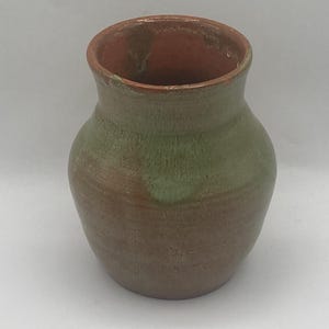 May include: A handmade ceramic vase with a rounded body and a slightly flared rim. The vase features a color gradient, transitioning from a warm brown at the base to a green midsection and a reddish-brown interior. The vase is suitable for displaying flowers.